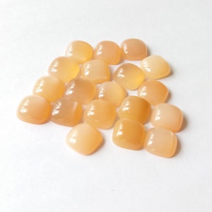May include: A pile of 18 smooth, light orange, square-shaped gemstones. The stones are arranged in a loose pile on a white background.