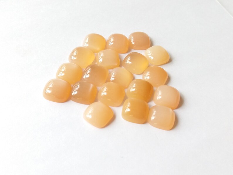 May include: A pile of 18 smooth, light orange, square-shaped gemstones. The stones are arranged in a loose pile on a white background.