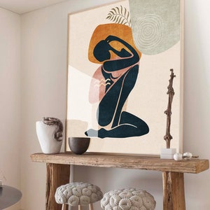 May include: A wooden console table with a rustic finish, featuring two round, tufted, gray poufs. The table is decorated with a large abstract print of a woman in a seated pose, with a vase and bowl on the surface.