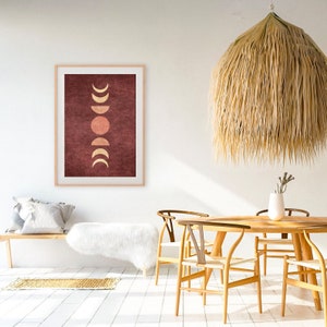 May include: A framed print with a minimalist design featuring a series of gold and peach moons and a sun on a burgundy background. The print is hanging on a white wall in a room with a wooden table and chairs.