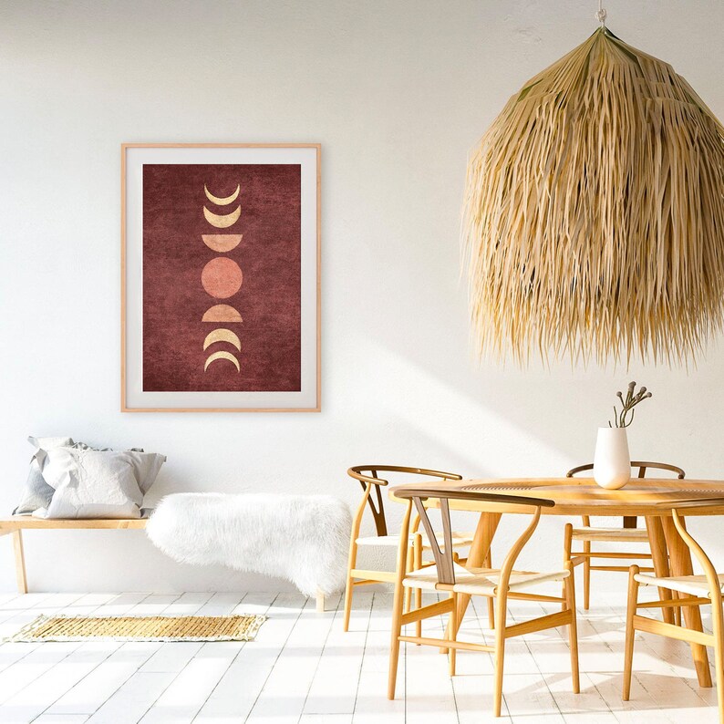 May include: A framed print with a minimalist design featuring a series of gold and peach moons and a sun on a burgundy background. The print is hanging on a white wall in a room with a wooden table and chairs.
