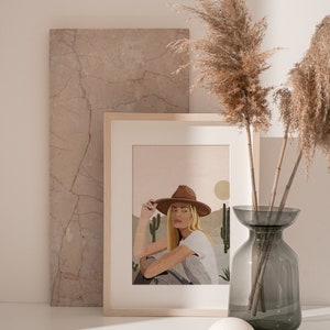 Puede incluir: Una ilustraci&oacute;n enmarcada de una mujer con un sombrero marr&oacute;n y una camisa blanca, sentada en un paisaje des&eacute;rtico con cactus. El fondo es un degradado suave rosa y naranja.