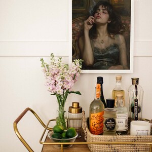 May include: A gold bar cart with a wooden top and a wicker basket filled with various bottles and a candle. The bottles include a clear bottle of ginger beer, a clear bottle of gin, a dark bottle of Domaine Canton, and a clear bottle of St-Germain. There are also limes in a glass bowl on the cart.