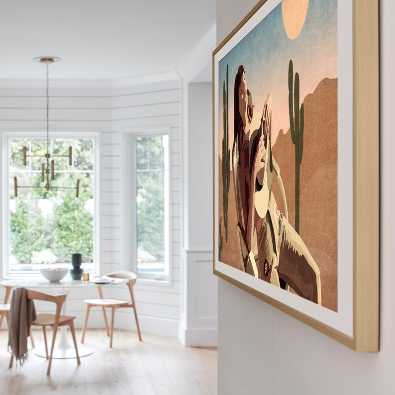 May include: A framed art print depicting a desert scene with a figure, cacti, and a sun. The artwork has a warm color palette of browns, tans, and blues. The frame is a light wood color, and the print is mounted on a white wall.