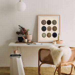 May include: A framed print of nine abstract circles in various shades of brown, beige, and blue. The print is hanging on a white wall above a desk with a white ceramic vase, a white ceramic mug, and two books.