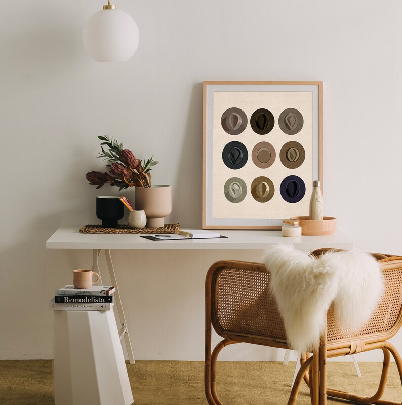 May include: A framed print of nine abstract circles in various shades of brown, beige, and blue. The print is hanging on a white wall above a desk with a white ceramic vase, a white ceramic mug, and two books.