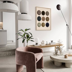 May include: A framed print of a minimalist art piece featuring nine circles with different colored hats inside each circle. The print is hanging on a white wall in a modern living room with a pink velvet armchair, a wooden bench, and a concrete coffee table.