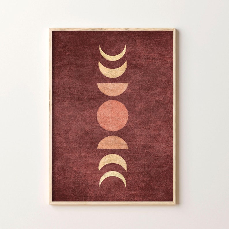 May include: A digital art print featuring a stylized moon phases design in gold and peach on a burgundy background. The print is framed in a light wood frame.