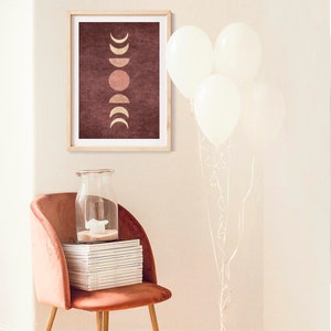 May include: A framed print with a maroon background and a gold moon phases design. The print is hanging on a white wall above a pink velvet chair with a stack of magazines and a glass jar.