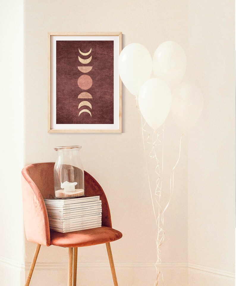 May include: A framed print with a maroon background and a gold moon phases design. The print is hanging on a white wall above a pink velvet chair with a stack of magazines and a glass jar.