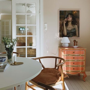 May include: A white table with a brown chair and a white lamp on a pink dresser with gold accents. A framed painting of a woman with dark hair and a black dress is hanging on the wall. The painting has the text "BOHO NEW ART" written on it.