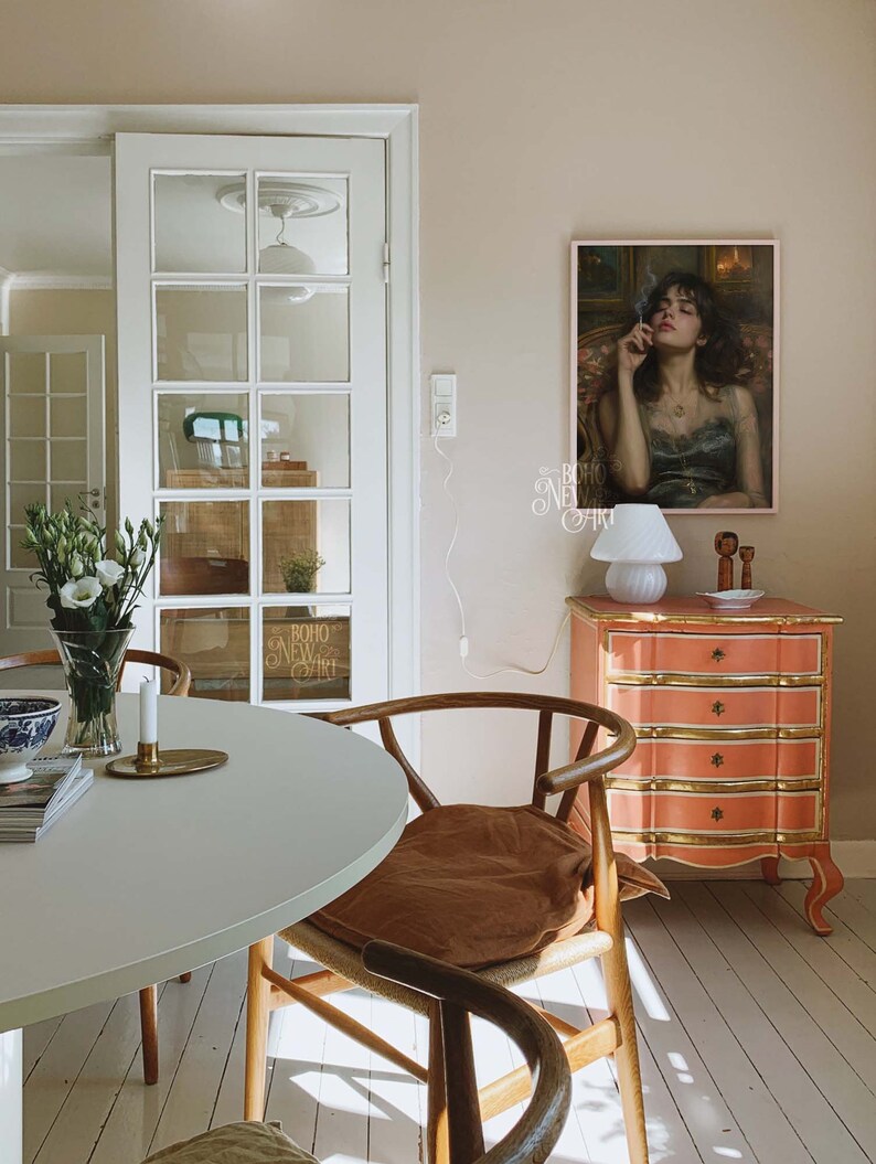 May include: A white table with a brown chair and a white lamp on a pink dresser with gold accents. A framed painting of a woman with dark hair and a black dress is hanging on the wall. The painting has the text "BOHO NEW ART" written on it.
