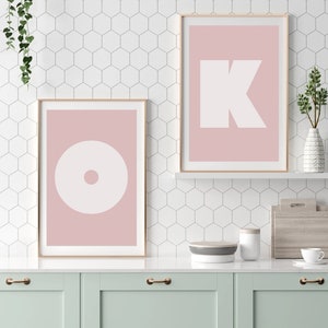 May include: Two framed prints with pink backgrounds and white geometric shapes. The print on the left has a circle with a smaller circle in the center. The print on the right has a large letter K.