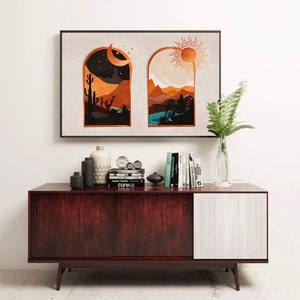 Op de afbeelding: Een ingelijst drukwerk met twee ramen, een toont een woestijnlandschap met een halve maan en de andere een bergketen met een zon. De print hangt aan een witte muur boven een donkerhouten kast met een witte deur.
