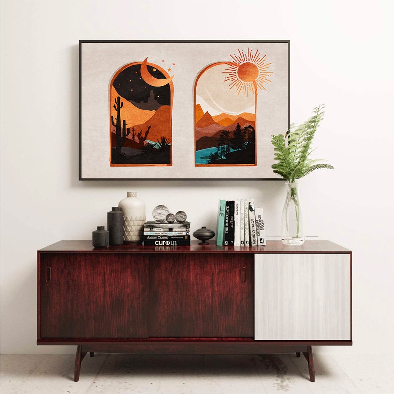 Op de afbeelding: Een ingelijst drukwerk met twee ramen, een toont een woestijnlandschap met een halve maan en de andere een bergketen met een zon. De print hangt aan een witte muur boven een donkerhouten kast met een witte deur.