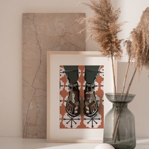May include: A framed art print featuring a stylized illustration of feet wearing brown sandals with decorative anklets. The sandals have a geometric design. The background shows a patterned orange and white tile floor.