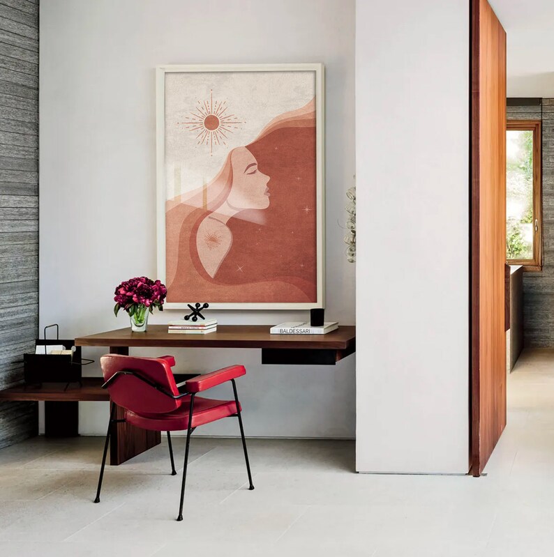 Puede incluir: Una impresi&oacute;n de arte minimalista que presenta el rostro de una mujer con el pelo suelto y un s&iacute;mbolo solar. La impresi&oacute;n est&aacute; en tonos de rojo, naranja y blanco y est&aacute; enmarcada en un marco blanco. La impresi&oacute;n est&aacute; colgada en una pared blanca sobre un escritorio de madera con una silla roja.