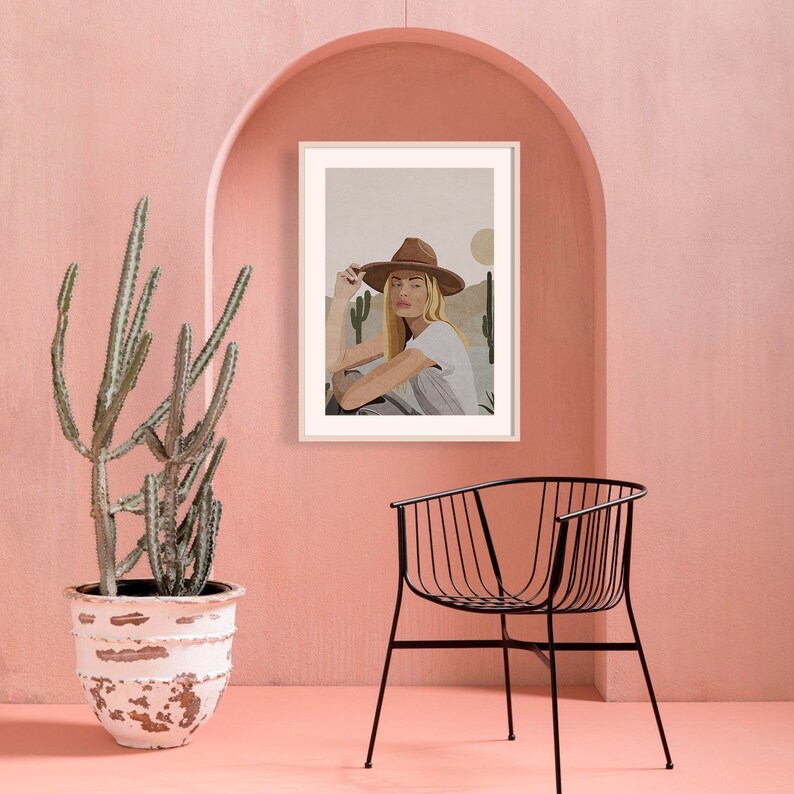 Puede incluir: Una impresi&oacute;n enmarcada de una mujer con un sombrero de vaquero marr&oacute;n y una camisa blanca, sentada en un paisaje des&eacute;rtico con cactus. La impresi&oacute;n est&aacute; colgada en una pared rosa.