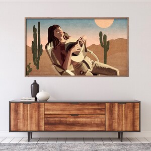 May include: A framed art print depicts a person relaxing in a chair under a desert sun. The artwork features a color palette of tan, brown, and blue, with cacti and a sun in the background. The person is wearing a hat and light-colored clothing.