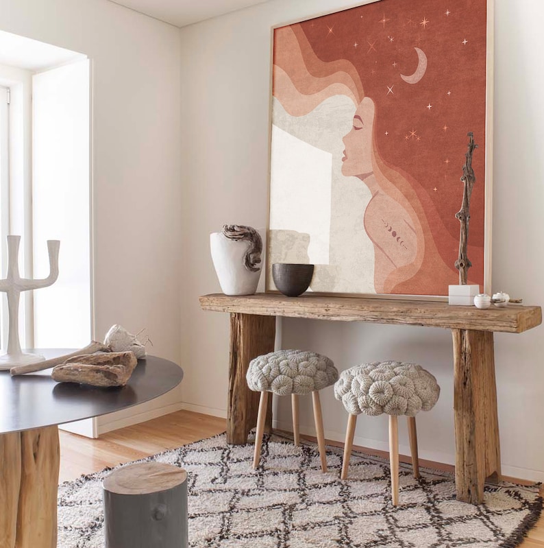 Puede incluir: Una mesa consola de madera r&uacute;stica con dos pufs de punto gris. La mesa tiene un acabado de madera natural y est&aacute; decorada con una gran pintura abstracta de una mujer con el pelo suelto y una luna creciente. La pintura tiene una combinaci&oacute;n de colores rojo, naranja y blanco.