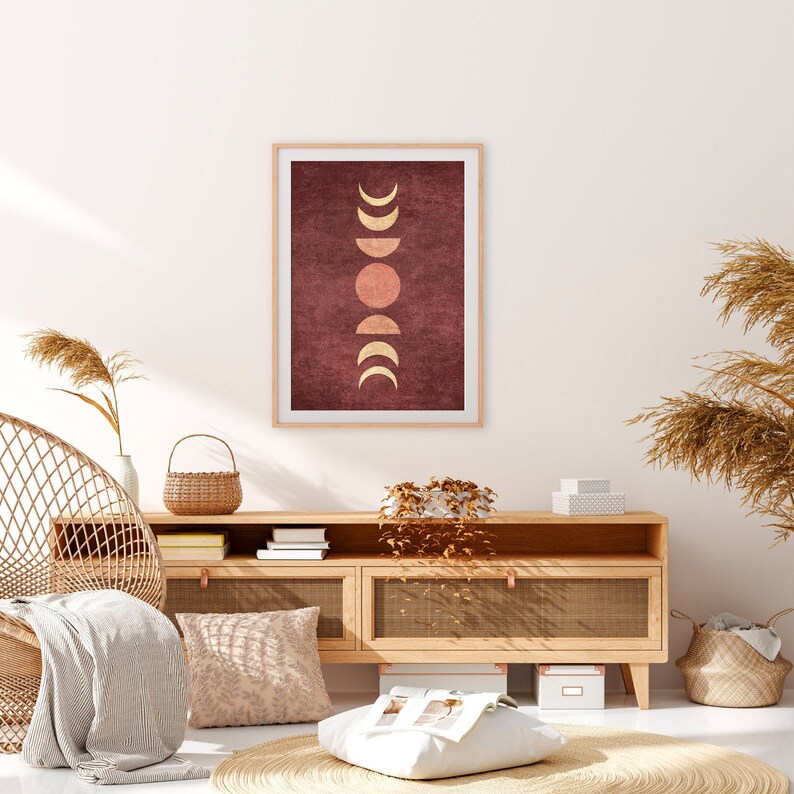 May include: A framed print with a minimalist design featuring a series of gold moons and a sun on a burgundy background. The print is hanging on a white wall above a wooden cabinet with shelves and drawers.