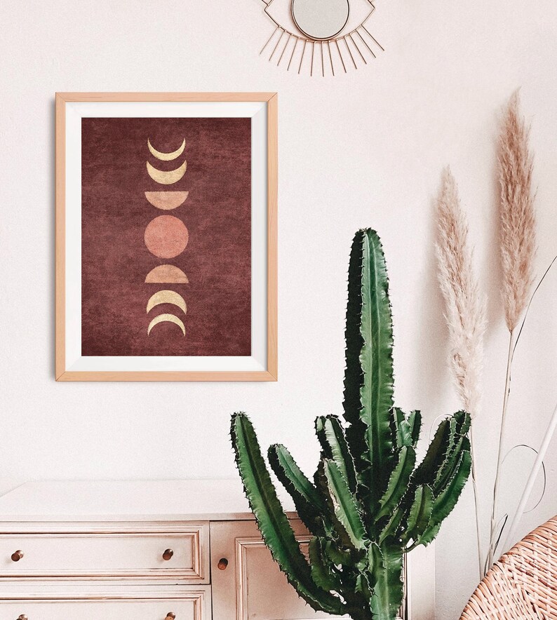 May include: A framed print with a maroon background and a gold moon phases design. The moon phases are in a vertical line, with a full moon in the center.