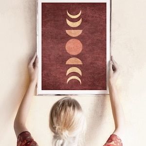 May include: A framed print with a minimalist design featuring a series of gold crescent moons and a red sun on a burgundy background.