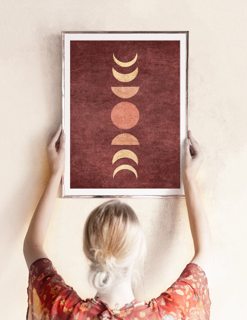 May include: A framed print with a minimalist design featuring a series of gold crescent moons and a red sun on a burgundy background.