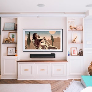 May include: A modern living room entertainment center with a large framed artwork depicting a desert scene. The room features built-in cabinets, shelves with decorative items, and a fireplace. A soundbar is mounted below the TV.
