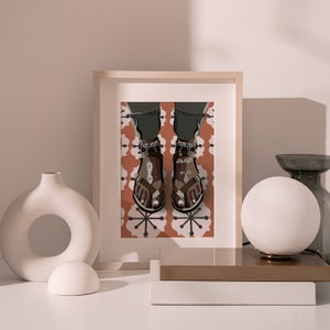 May include: An illustration of a framed artwork featuring a person's feet wearing brown sandals with silver anklets. The sandals have a geometric design. The background has a repeating floral pattern in shades of orange and white.