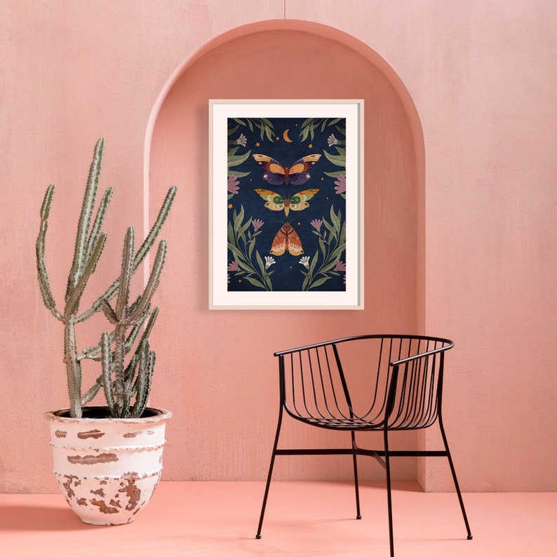 May include: A framed print with a dark blue background featuring three stylized moths with floral accents. The moths are a dark purple, green, and orange color. The print is surrounded by a light wood frame.