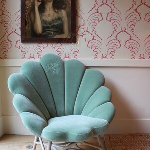 May include: A teal blue velvet chair with a shell-shaped back and white wicker legs. The chair is in front of a white wall with pink floral wallpaper. The chair is in a room with a wooden floor.