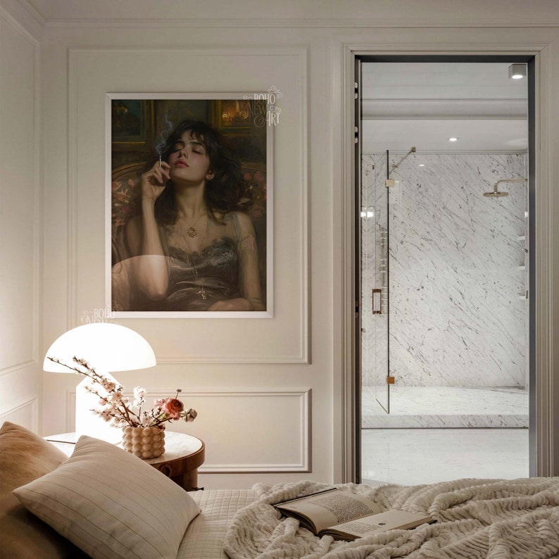 May include: A white lamp with a white shade sits on a wooden nightstand next to a bed with a white blanket and a book. A framed print of a woman with dark hair and a necklace hangs on the wall above the nightstand.