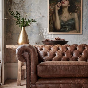 May include: A brown leather chesterfield sofa with button tufting sits in front of a wall with a framed painting of a woman in a blue dress holding a cigarette. The painting is set against a dark background with a small framed picture above it.