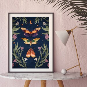 May include: A framed print with a dark blue background featuring three moths with intricate patterns and colors. The moths are surrounded by green leaves and pink flowers. The print has a crescent moon and stars in the top center.