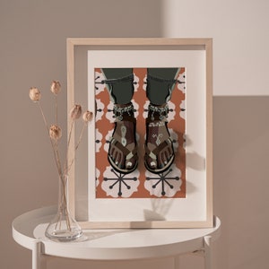May include: A framed art print featuring an illustration of feet wearing brown sandals with decorative ankle bracelets. The sandals have a geometric design. The background is a pattern of white flowers on an orange background.