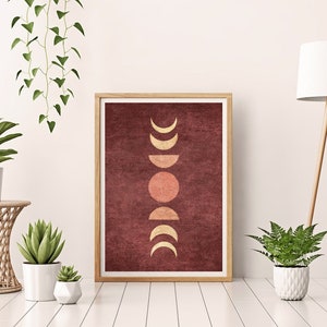 May include: A framed print with a minimalist design featuring a series of gold moons and a sun on a burgundy background. The print is displayed on a white wall with a potted plant and a floor lamp.