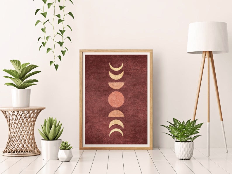 May include: A framed print with a minimalist design featuring a series of gold moons and a sun on a burgundy background. The print is displayed on a white wall with a potted plant and a floor lamp.