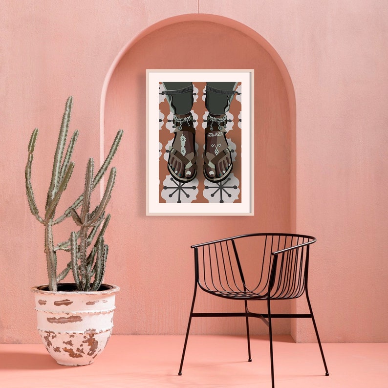 May include: An art print featuring a person's feet wearing brown sandals with silver anklets, set against a patterned background. The print is framed and hangs on a pink wall, with a cactus and black chair in the foreground.