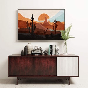 May include: A framed print of a desert landscape with a sunset in the background. The print features a silhouette of cacti and mountains. The print is hanging on a white wall above a dark wood cabinet with books and decorative objects.