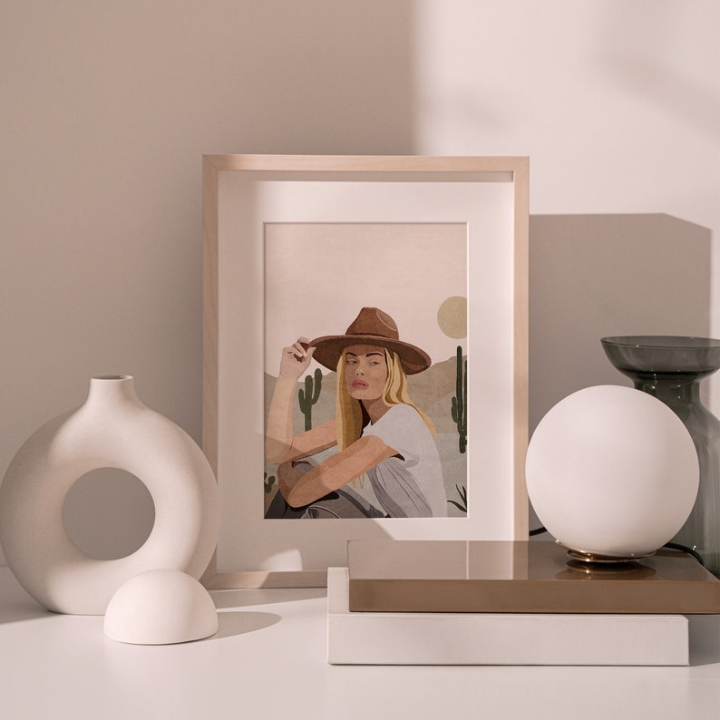 Puede incluir: Una impresi&oacute;n enmarcada de una mujer con un sombrero marr&oacute;n y una camisa blanca, sentada en un paisaje des&eacute;rtico con cactus. La impresi&oacute;n est&aacute; en un estante blanco con un jarr&oacute;n blanco y una l&aacute;mpara de globo blanco.