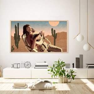 May include: A framed art print depicts a person relaxing in a chair in a desert landscape. The artwork features a warm color palette with a sun, cacti, and mountains. The person is wearing a hat and light-colored clothing.