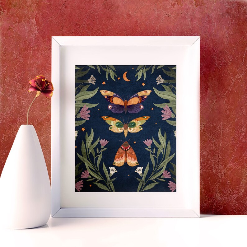 May include: A framed print with a dark blue background featuring three colorful moths with intricate patterns and a crescent moon. The moths are surrounded by green leaves and white flowers.