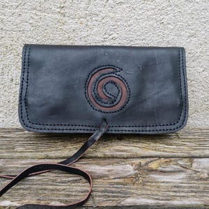 May include: Black leather clutch with a brown spiral design. The clutch has a flap closure and a long, thin strap. The edges are stitched with black thread. The bag is displayed on a wooden surface.