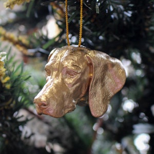 Könnte beinhalten: Ein goldfarbener Weihnachtsbaumschmuck in Form eines Hundekopfes. Der Schmuck hängt an einer goldenen Schnur an einem Weihnachtsbaum.