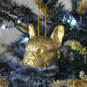 May include: A gold-colored French bulldog ornament hanging from a Christmas tree. The ornament is detailed and realistic, with a textured surface.