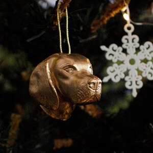 Puede incluir: Un adorno de cabeza de perro de color dorado que cuelga de un árbol de Navidad. El adorno es detallado y realista, con una superficie texturizada.