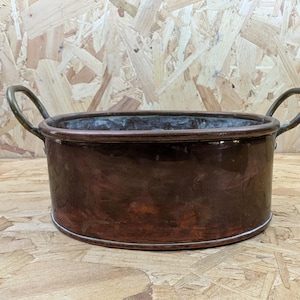 May include: A copper-colored, round planter with two brass handles. The planter has a dark, aged patina, and a bright, reflective rim. The handles curve upwards from the sides. The planter is suitable for indoor or outdoor use.