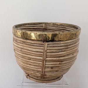 Könnte beinhalten: Ein runder Korbübertopf mit einem Korpus aus naturfarbenem, hellbraunem geflochtenem Rattan und einem glatten, polierten Messingrand an der Oberseite. Die Struktur besteht aus horizontalen Rattanstreben, die durch vertikale Stützen gehalten werden.