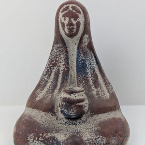 May include: A ceramic sculpture of a seated figure with a draped head covering. The figure is primarily brown with white and blue accents, and holds a small, round vessel. The sculpture has a textured surface and a serene expression.